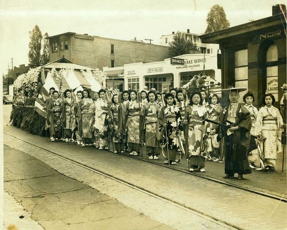 Two rows of several kimono dressed Japanese American women stand posing for the camera in front of a float decorated for the Narrows Bridge Parade on Jefferson Avenue, Tacoma, WA The heads of several men and women peer out of the float which is in photo left. A man in Japanese dress stands at photo right at the head of the rows. The women hold streamers in their hands, appearing as though they will be pulling the float. Street car tracks are on the street, and a brake repair business is behind the women. Kimi Tanbara stands beneath the name Rowland on the business front. The Northern Laundry business is at photo right. Yamane Family Collection. Sarah Sugimoto is second from right.