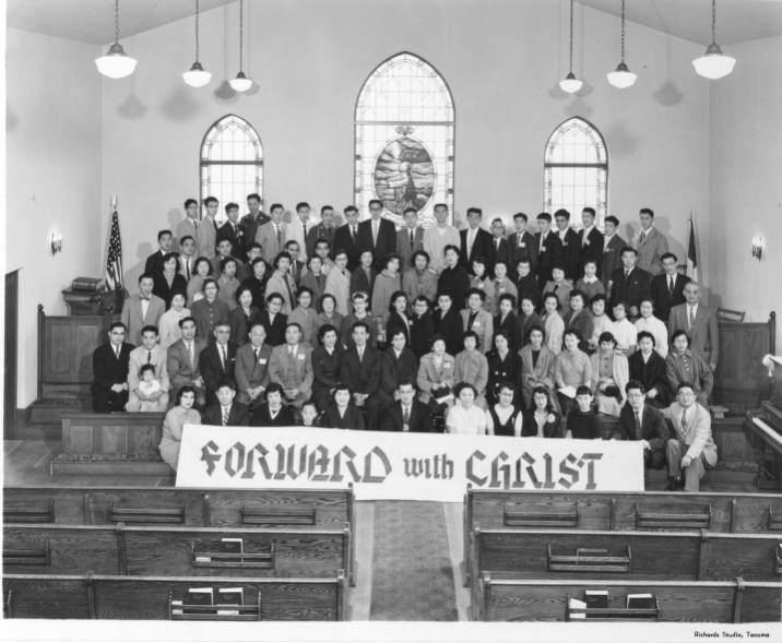 Large group of Japanese Americans inside a church. Wooden pews and a “Forward with Christ” sign are in front of the group. 