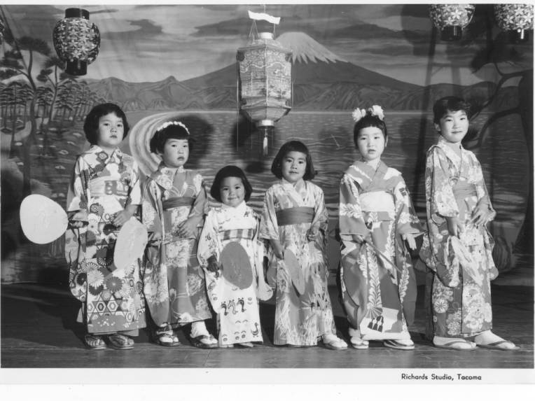 Group of six Japanese American girls in kimonos in front of a scenic stage backdrop.