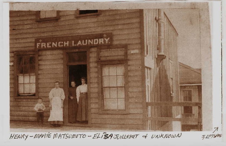 Sepia photo with a wooden building, two Japanese American children, and a White woman standing in a doorway.