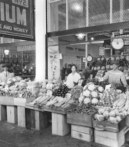 Black-and-white photo of a produce stand, with several Japanese American people standing behind tables of vegetables and fruits.