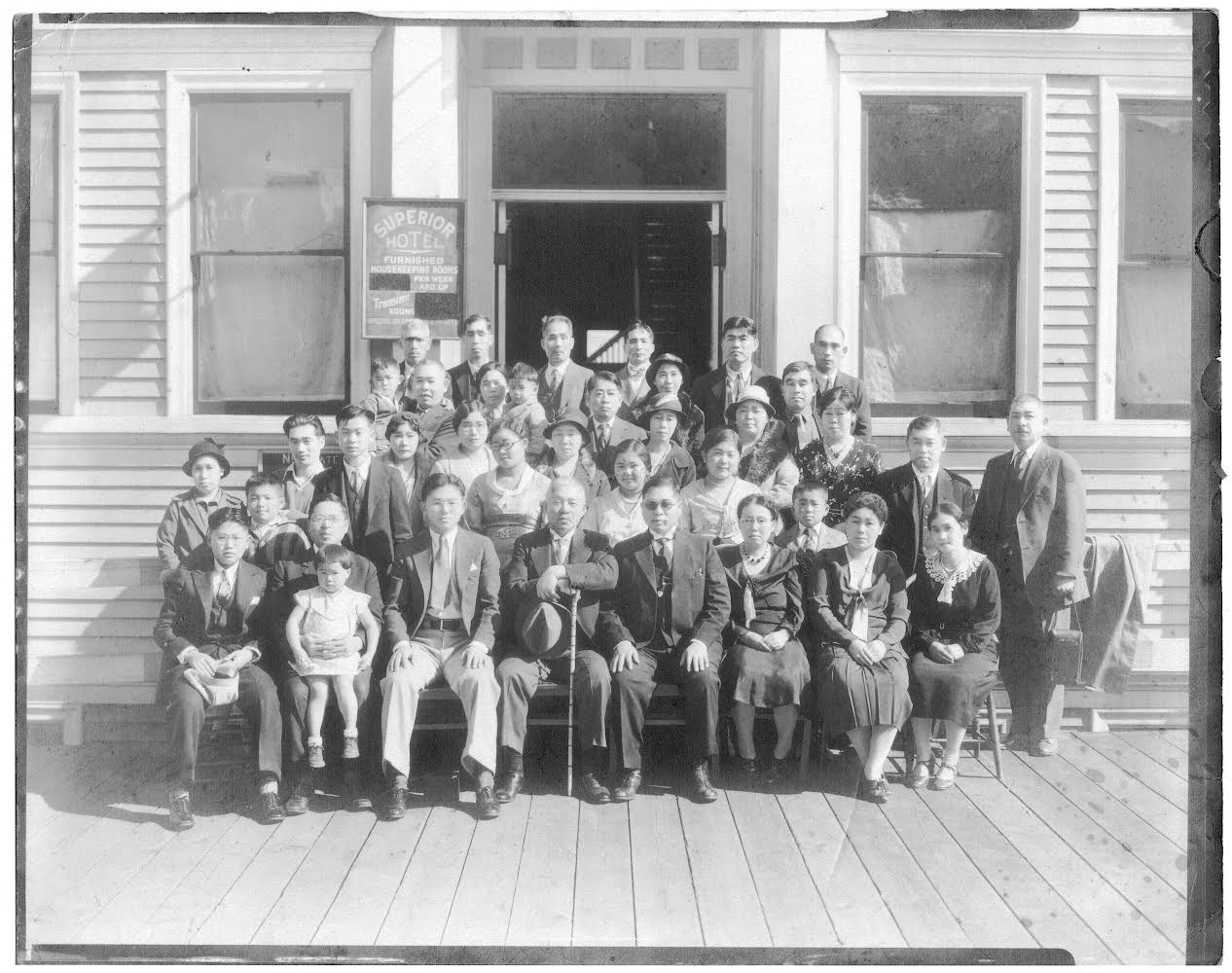 Black-and-white photo of a group of Japanese people in suits in front of a wooden hotel with windows.